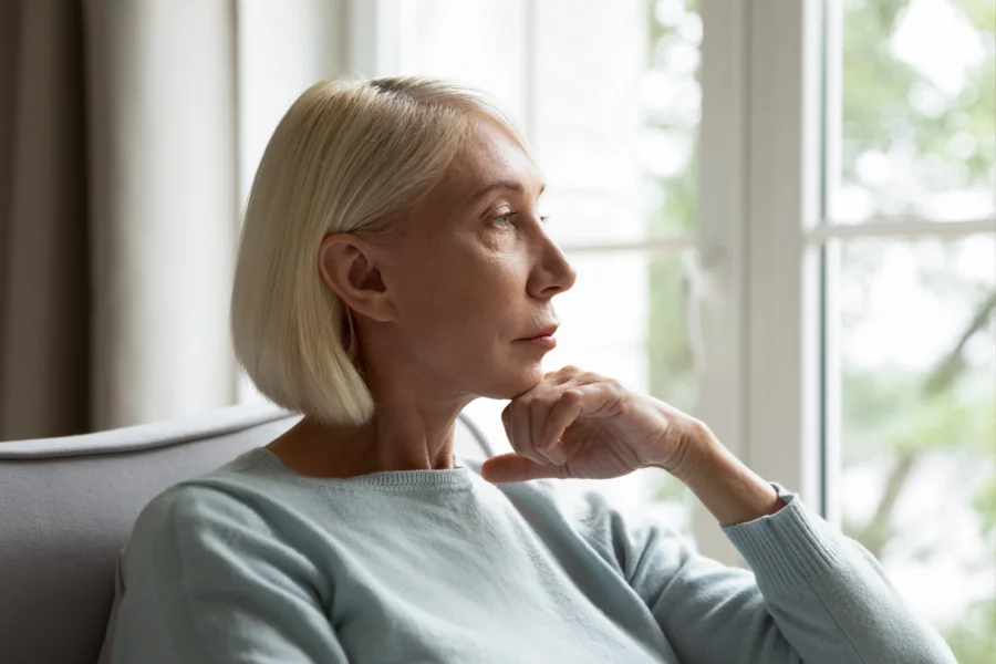 A middle-aged woman with silver hair sits in quiet contemplation by a window, reflecting on how long perimenopause lasts.