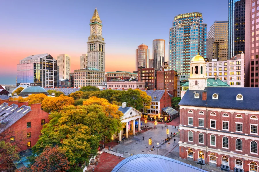 Boston’s downtown skyline and the Faneuil Hall marketplace glow at sunset above autumn trees.