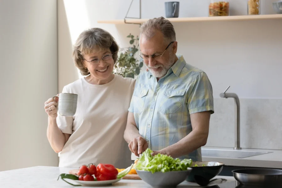 A smiling older couple prepares a fresh vegetable salad together in a bright home kitchen while discussing gut microbiome testing.
