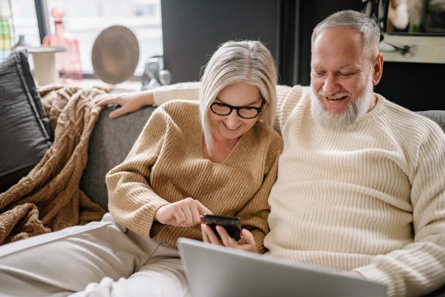 An older couple in cozy sweaters relaxes on a couch, viewing GLP-1 patch information on a phone.